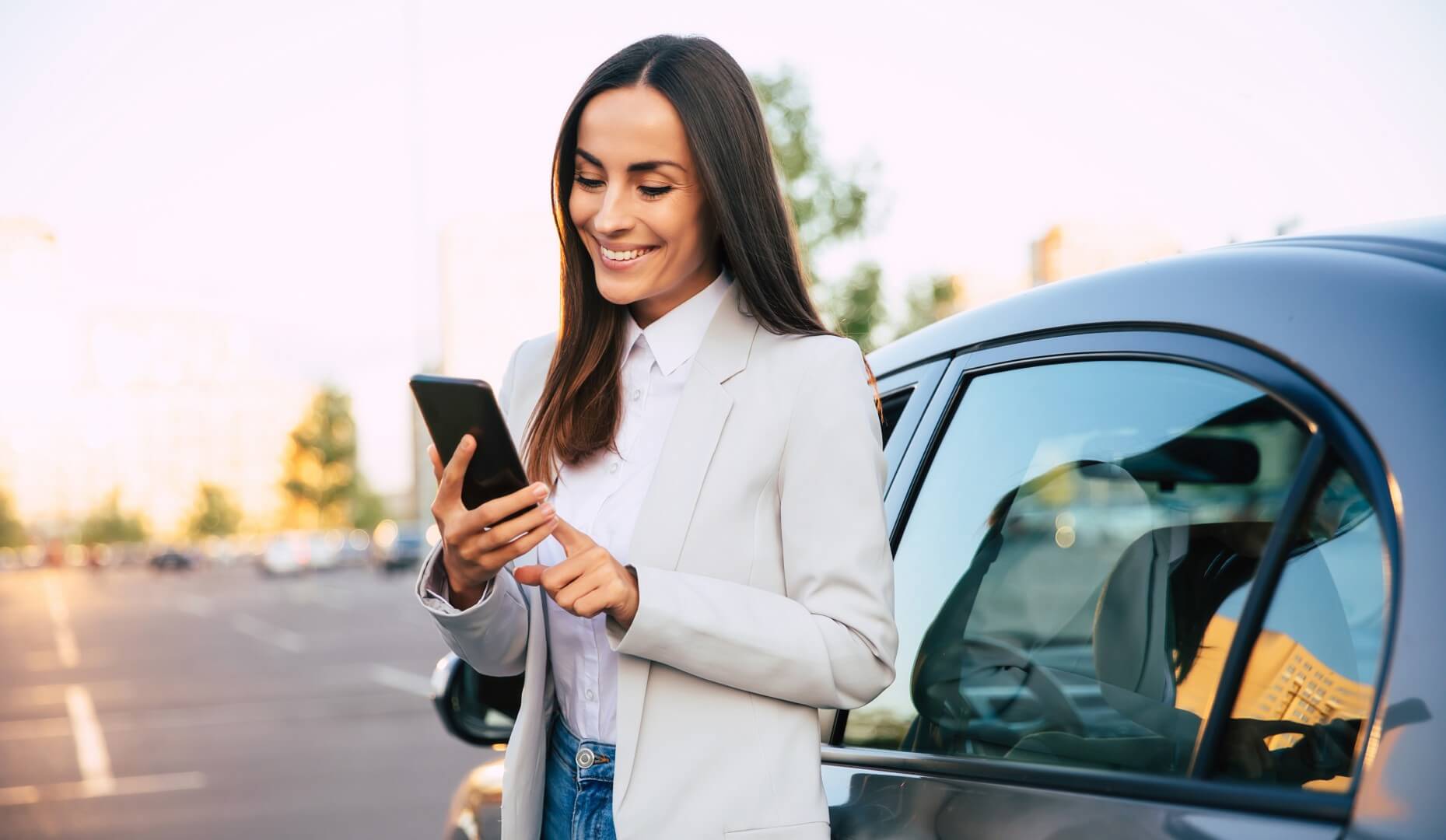 Geschäftsfrau in hellem Blazer steht lächelnd neben ihrem Auto auf einem sonnigen Parkplatz und schaut auf ihr Smartphone.